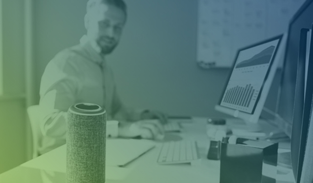 Homem em sua mesa trabalhando no computador e ouvindo um smart speaker