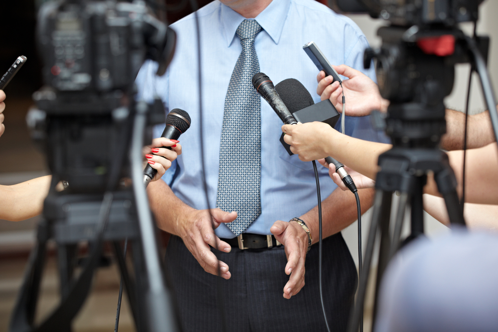Homem com trajes sociais falando em microfones em frente de uma câmera