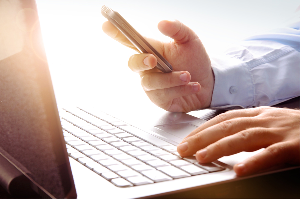 Homem utilizando computador com celular na mão