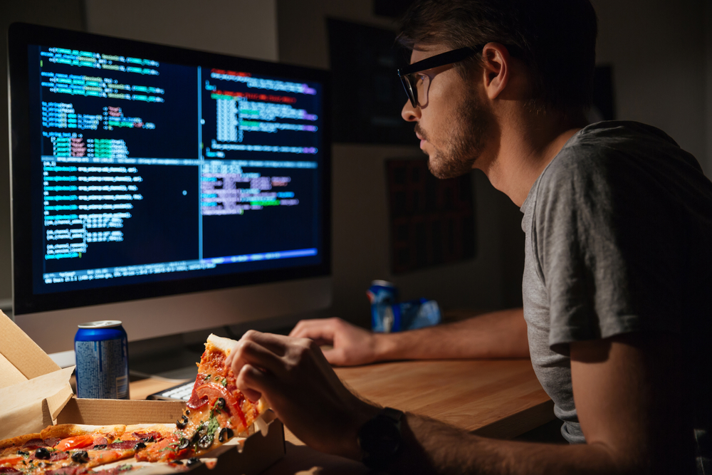 Homem comendo pizza e utilizando computador com tela exibindo código