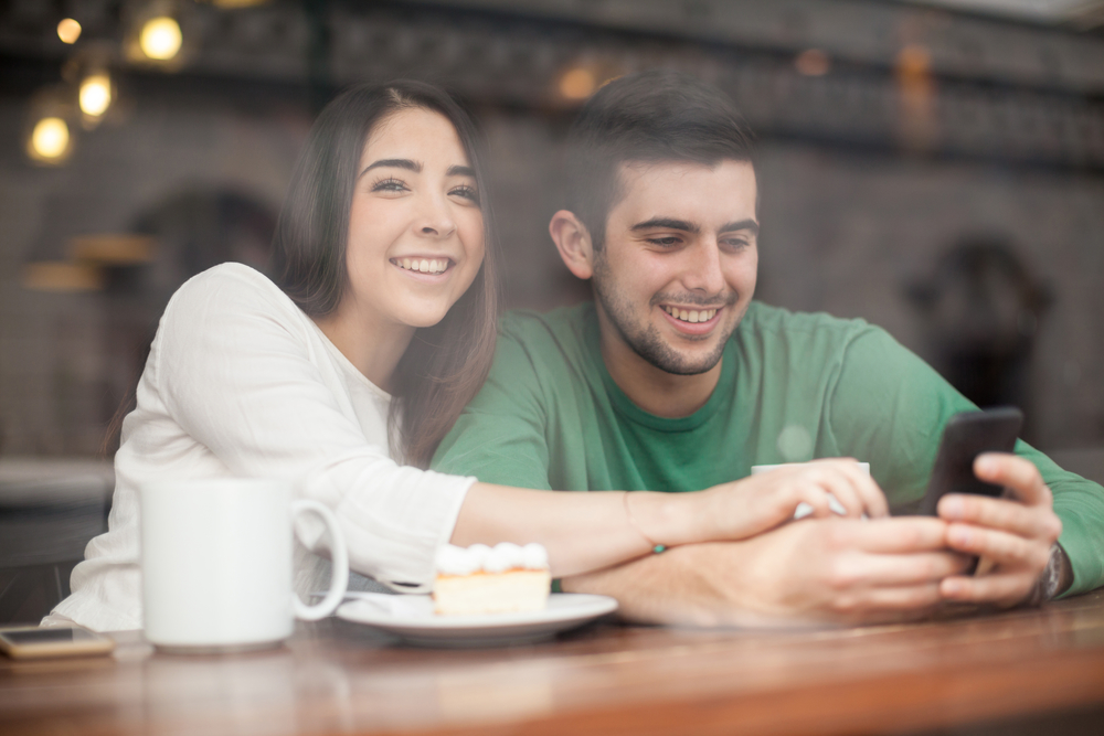 Homem e mulher sorrindo e usando o celular dentro de um café